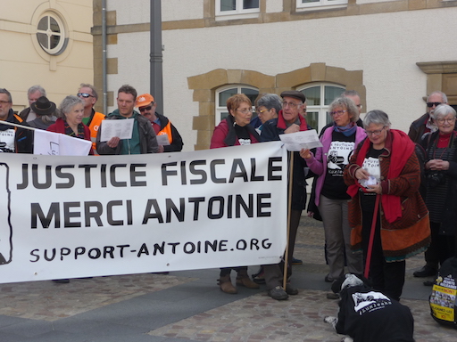 Des membres du Comité de soutien chantent groupés derrière une banderolle “Justice Fiscale, merci Antoine!”