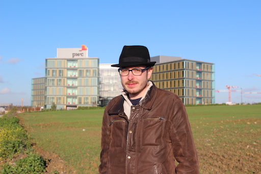 Antoine Deltour, debout dans un champ, chapeau sur la tête. Au fond, le bâtiment principal de pwc Luxembourg.